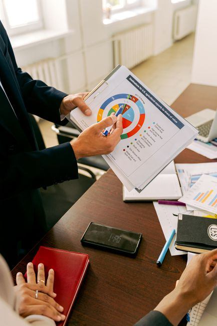 A person holds a printed chart with colorful data circles while standing at a desk covered with papers, a phone, and a notebook.