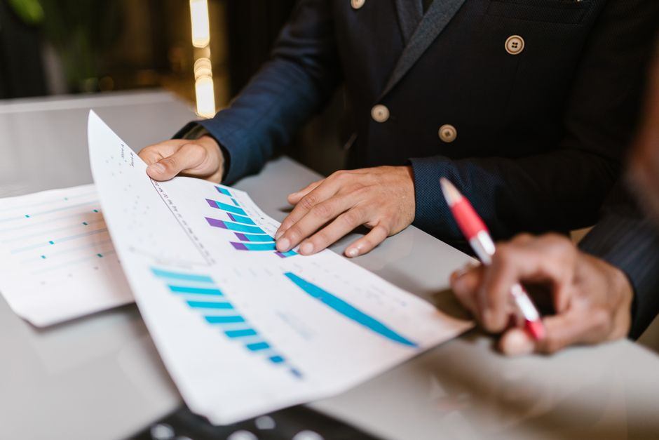 A person in a suit points to a printed bar chart on a desk while another holds a pen nearby.